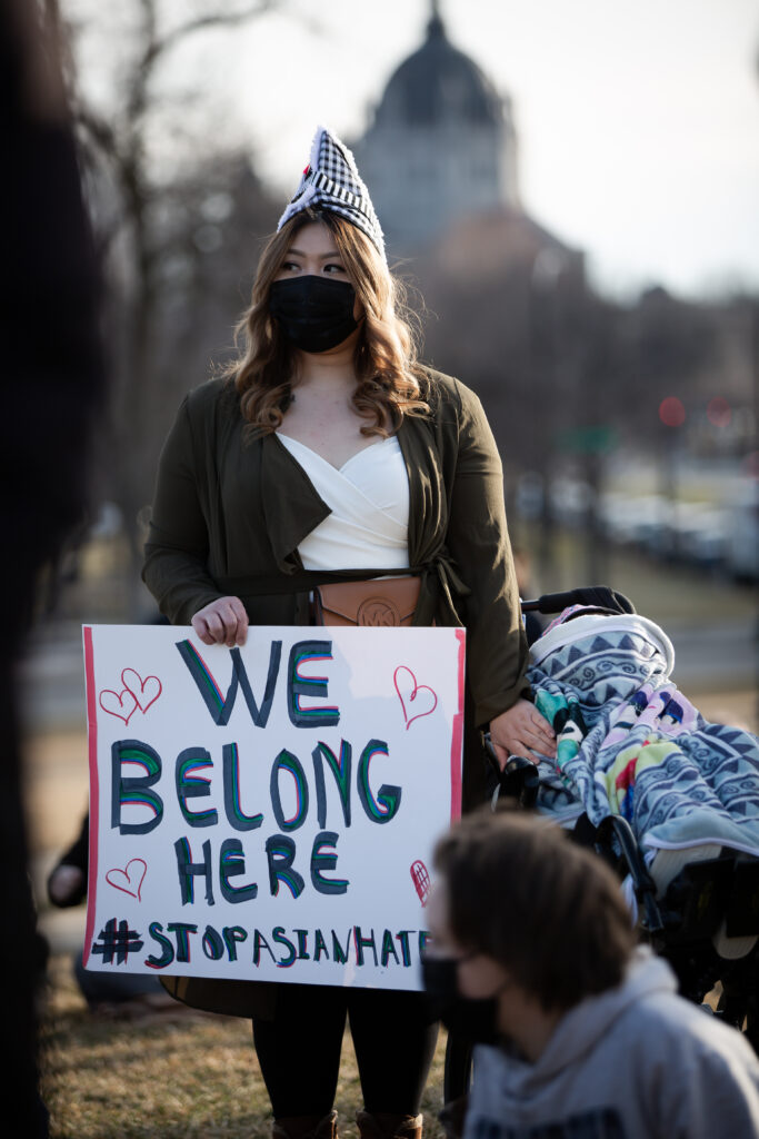 A young woman in a mask at the Minnesota Capitol grounds holding a sign that says We Belong Here