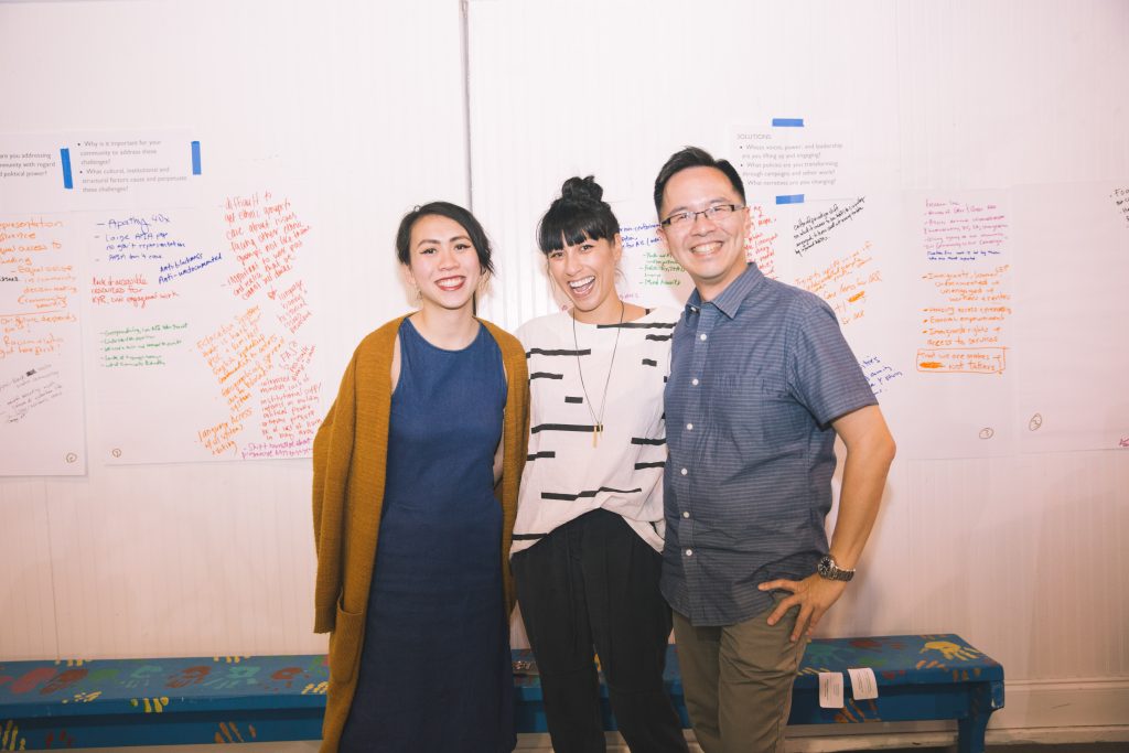 Three people stand, posing for a camera. Behind them is a wall with taped papers with writing.