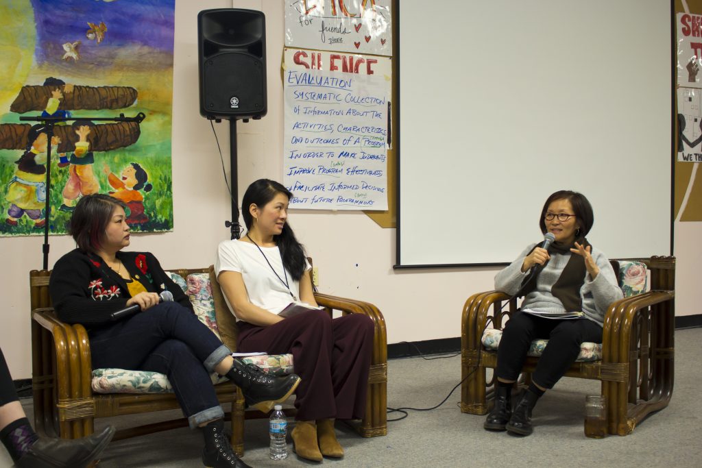 Three women are seated. Two sit on the left, sharing a couch, while one sits on the right in an armchair, holding a microphone as she speaks.