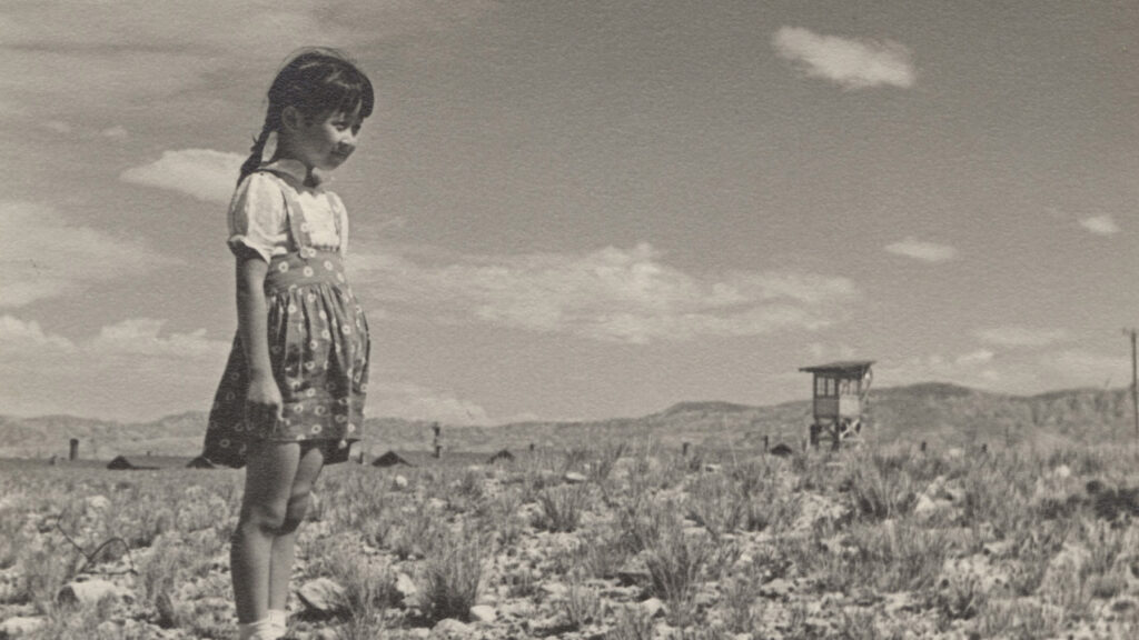 Photo entitled "Okumoto Girl with Guard Tower". A little girl looks across a barren field with a guard tower in the background.